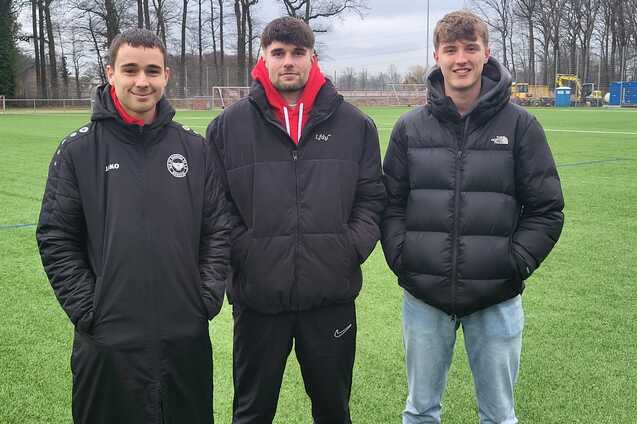 Drei junge M&auml;nner stehen auf einem Fu&szlig;ballplatz, umgeben von B&auml;umen, mit Sportjacken und verschiedenen Outfits.