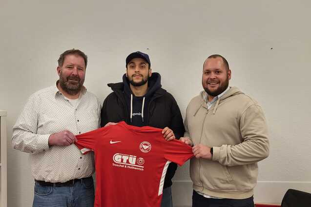 Drei M&auml;nner halten gemeinsam ein rotes Fu&szlig;balltrikot mit Logo, vor einem neutralen Hintergrund.