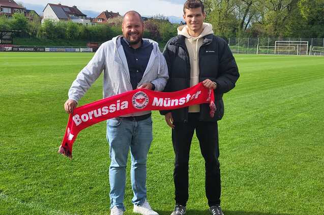 Zwei M&auml;nner stehen auf einem Fu&szlig;ballplatz und halten einen roten Schal mit der Aufschrift "Borussia M&uuml;nster".