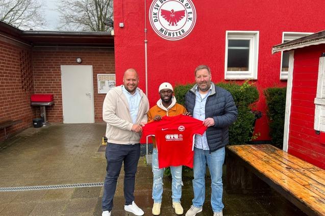 Drei M&auml;nner stehen vor einem Vereinsgeb&auml;ude und halten ein rotes Trikot des Fu&szlig;ballvereins Borussia M&uuml;nster.