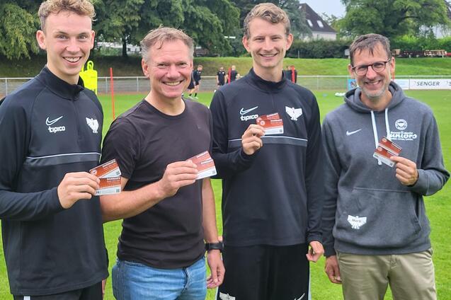Vier M&auml;nner halten stolz ihre Mitgliedskarten vor einem Fu&szlig;ballfeld, umgeben von B&auml;umen und einem Sportplatz.