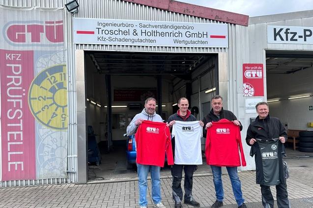 Vier M&auml;nner stehen vor einem GT&Uuml;-Geb&auml;ude und halten T-Shirts in den Farben Rot und Wei&szlig; sowie eine schwarze Tasche.