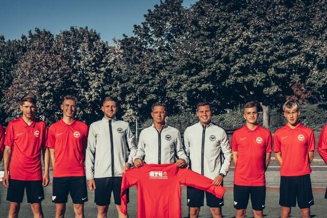 Teamfoto von acht Fu&szlig;ballspielern in roten und grauen Trikots, auf einem Sportplatz mit B&auml;umen im Hintergrund.