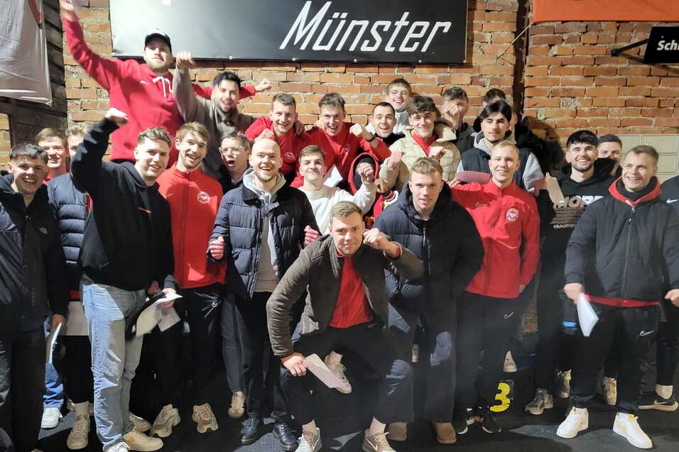 Gruppennfoto einer Fu&szlig;ballmannschaft in roten Trikots, l&auml;chelnd und posierend in einer Indoor-Location.