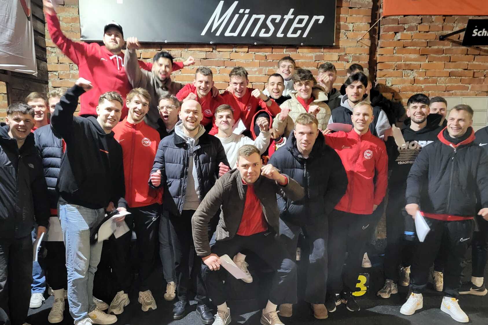 Gruppennfoto einer Fu&szlig;ballmannschaft in roten Trikots, l&auml;chelnd und posierend in einer Indoor-Location.