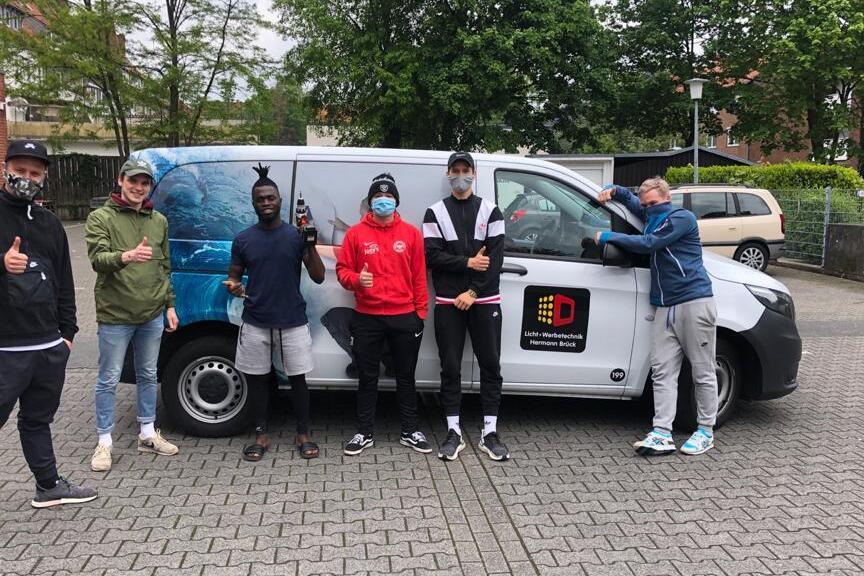 Sechs Personen posieren vor einem Lieferwagen, alle zeigen Daumen hoch und tragen Masken. Im Hintergrund sind B&auml;ume sichtbar.