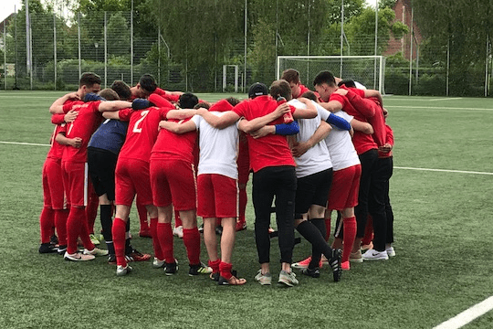 Eine Gruppe von Fu&szlig;ballspielern in roten Trikots umarmt sich auf einem Sportplatz im Kreis.