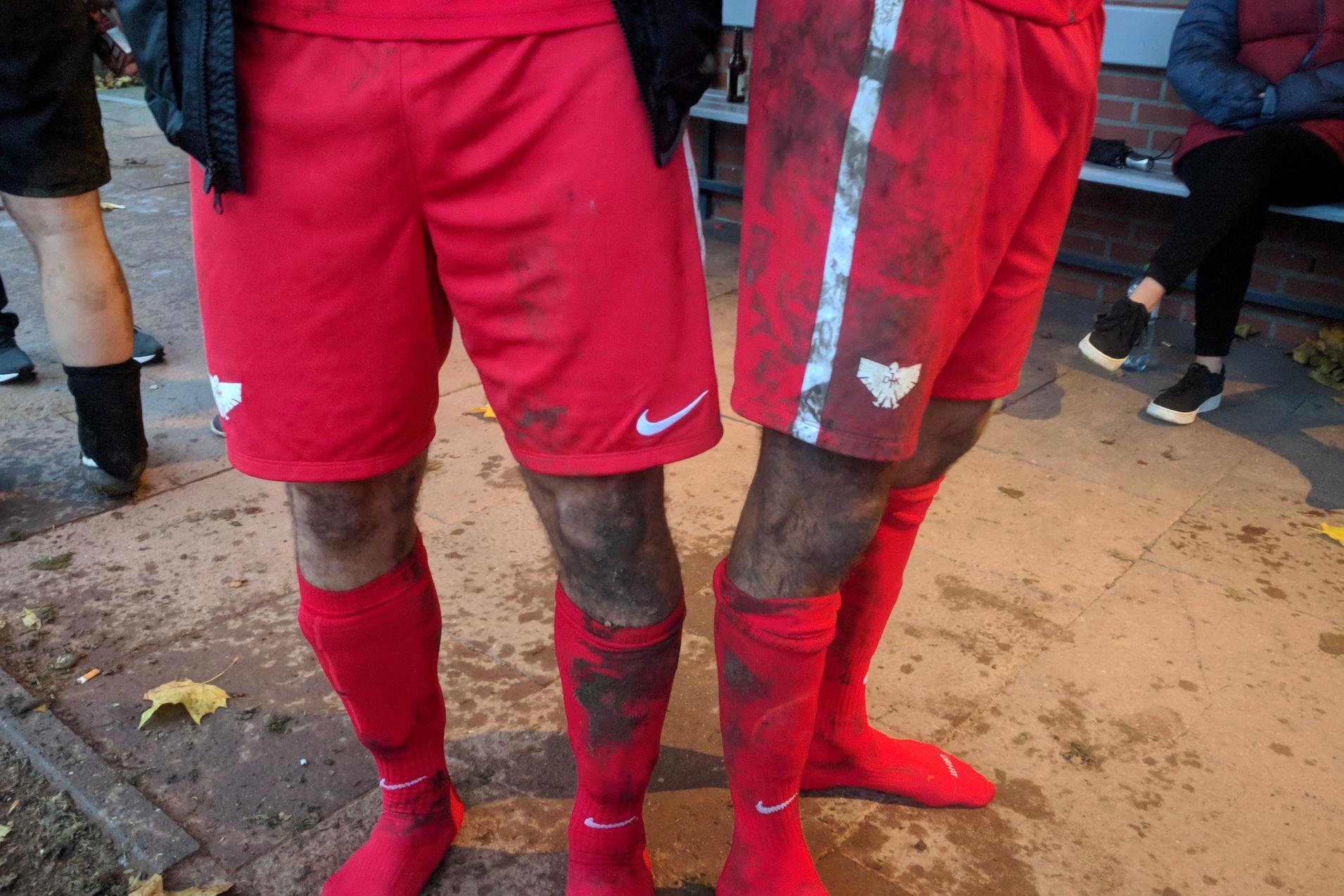 Zwei Spieler in roten Fu&szlig;balltrikots mit schmutzigen Hosen und Socken stehen auf einem Platz. Im Hintergrund sind Zuschauer.