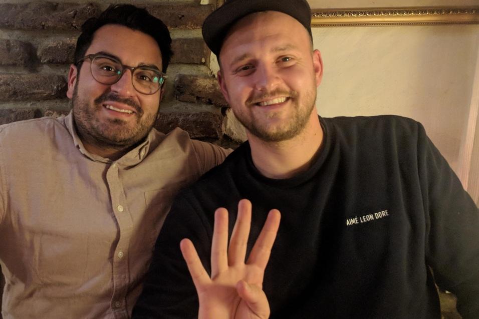 Zwei M&auml;nner sitzen an einem Tisch, einer zeigt mit der Hand die Zahl vier, beide l&auml;cheln in die Kamera.