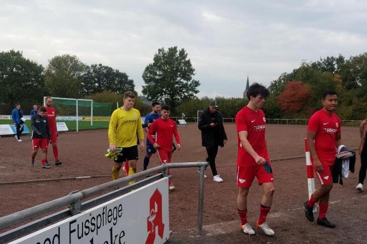 Fu&szlig;ballmannschaft in roten Trikots verl&auml;sst das Spielfeld, Zuschauer stehen am Rand des Platzes. Wolkiger Himmel.