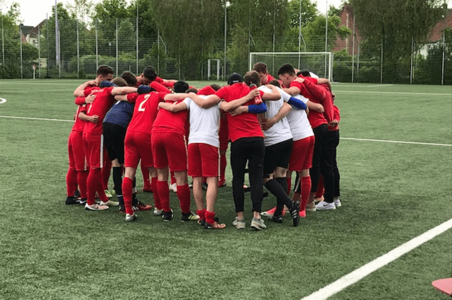 Eine Gruppe von Fu&szlig;ballspielern in roten Trikots umarmt sich auf einem Sportplatz im Kreis.