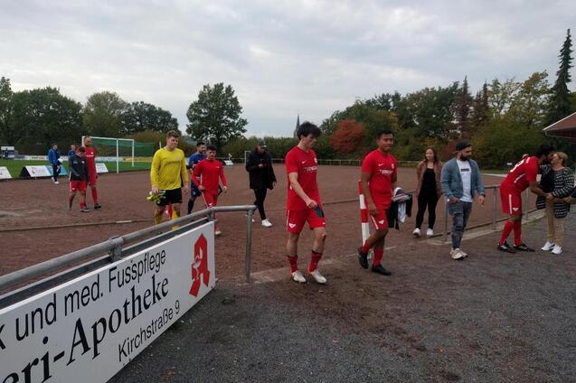 Fu&szlig;ballmannschaft in roten Trikots verl&auml;sst das Spielfeld, Zuschauer stehen am Rand des Platzes. Wolkiger Himmel.