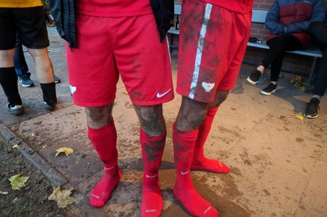 Zwei Spieler in roten Fu&szlig;balltrikots mit schmutzigen Hosen und Socken stehen auf einem Platz. Im Hintergrund sind Zuschauer.