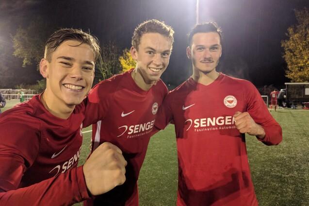 Drei junge Fu&szlig;ballspieler in roten Trikots posieren l&auml;chelnd auf einem beleuchteten Platz bei Nacht.