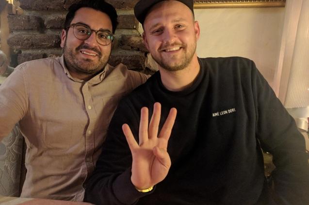 Zwei M&auml;nner sitzen an einem Tisch, einer zeigt mit der Hand die Zahl vier, beide l&auml;cheln in die Kamera.