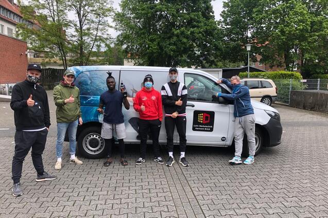 Sechs Personen posieren vor einem Lieferwagen, alle zeigen Daumen hoch und tragen Masken. Im Hintergrund sind B&auml;ume sichtbar.