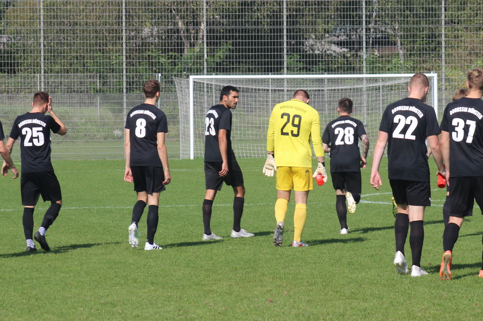 Acht Fu&szlig;ballspieler in schwarzen Trikots laufen auf ein Tor zu, ein Torwart in gelben Trikot geht voraus.