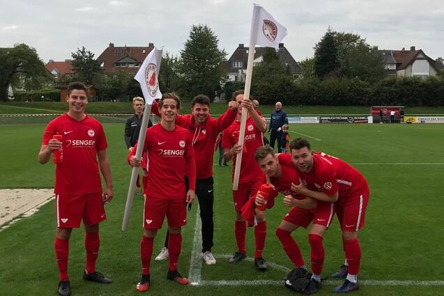 Sechs Fu&szlig;ballspieler in roten Trikots posieren l&auml;chelnd mit Fahnen auf einem gr&uuml;nen Fu&szlig;ballplatz.