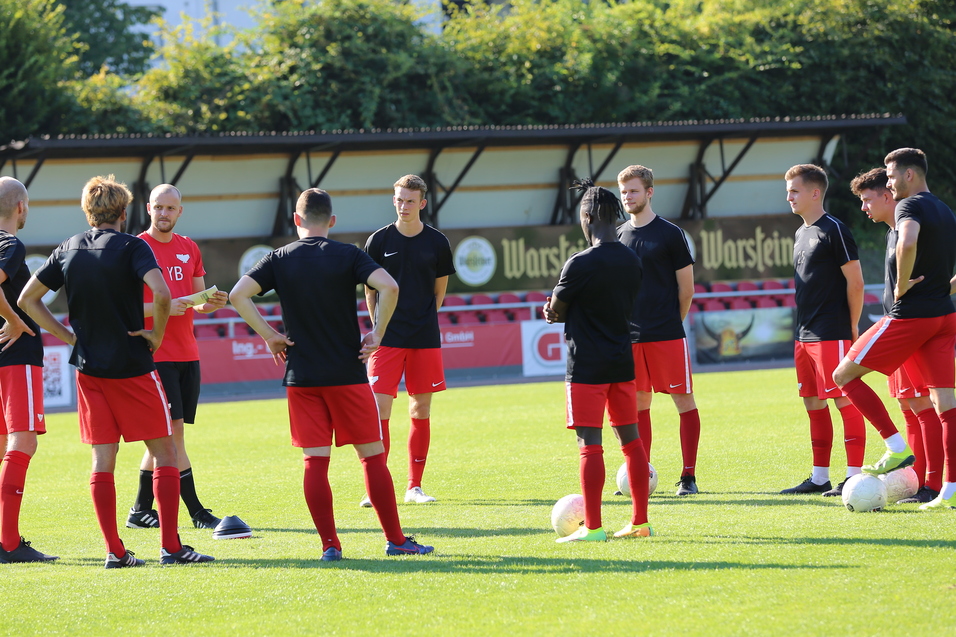 Eine Gruppe von neun Fu&szlig;ballspielern in schwarzer und roter Kleidung steht auf einem Rasenplatz und trainiert.