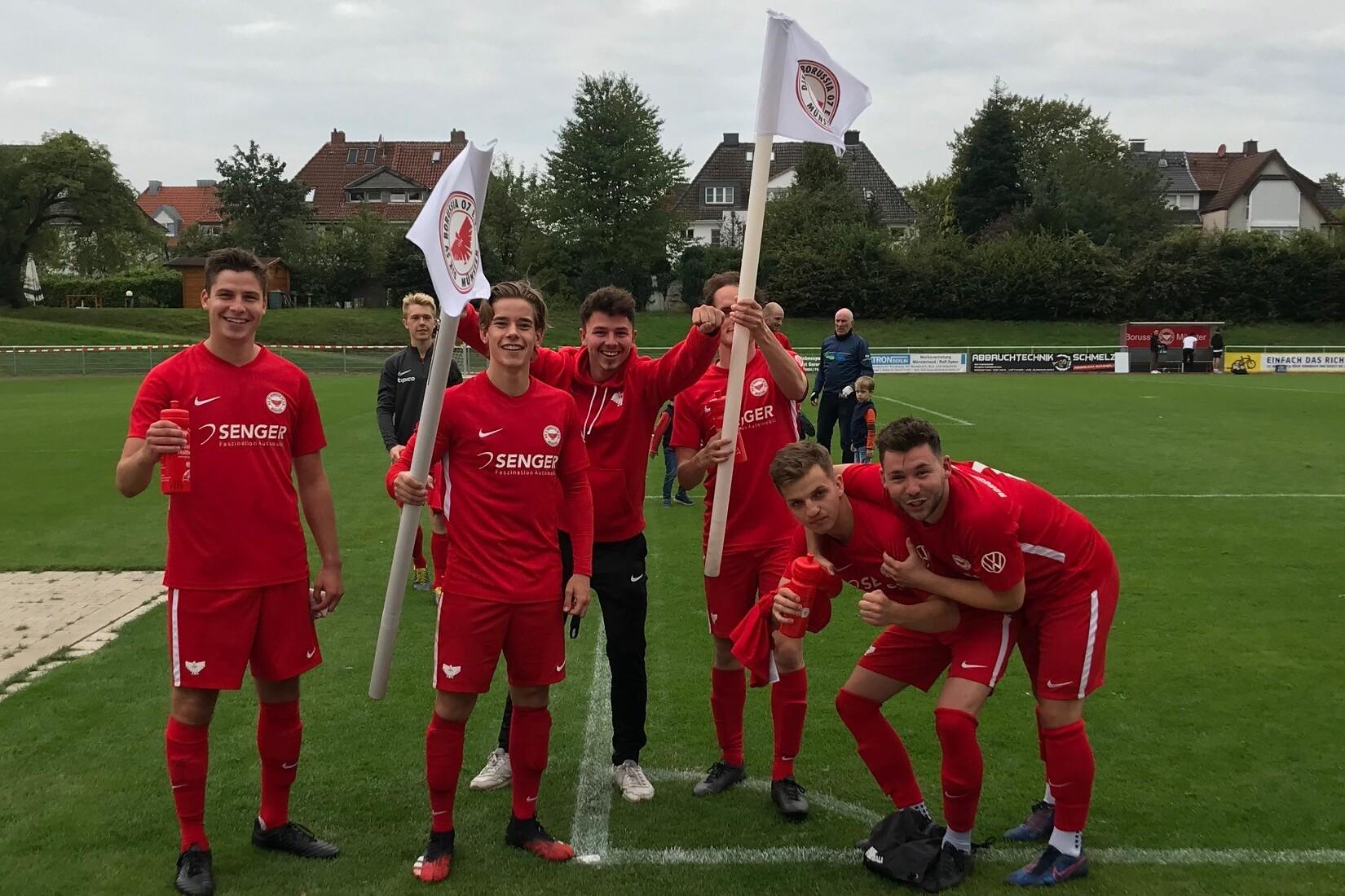 Sechs Fu&szlig;ballspieler in roten Trikots posieren l&auml;chelnd mit Fahnen auf einem gr&uuml;nen Fu&szlig;ballplatz.