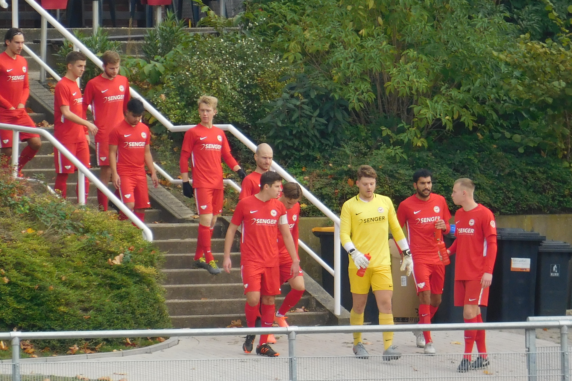 Fu&szlig;ballmannschaft in roten Trikots betritt das Spielfeld, gefolgt von Schiedsrichtern in gelben Trikots.