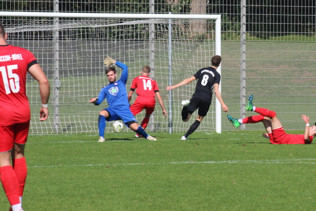 Spielszene auf einem Fu&szlig;ballfeld: Ein Spieler in Rot f&auml;llt, w&auml;hrend ein Torh&uuml;ter in Blau den Ball abwehrt.