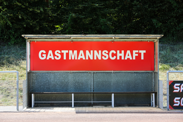 Sitzbank f&uuml;r die Gastmannschaft mit rotem Schild auf einem Sportplatz.