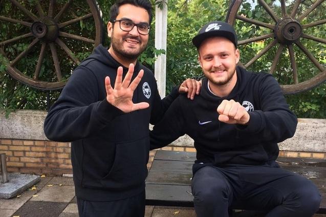 Zwei M&auml;nner in schwarzen Hoodies posieren freundlich vor einem Rad und einer gr&uuml;nen Pflanzenwand.