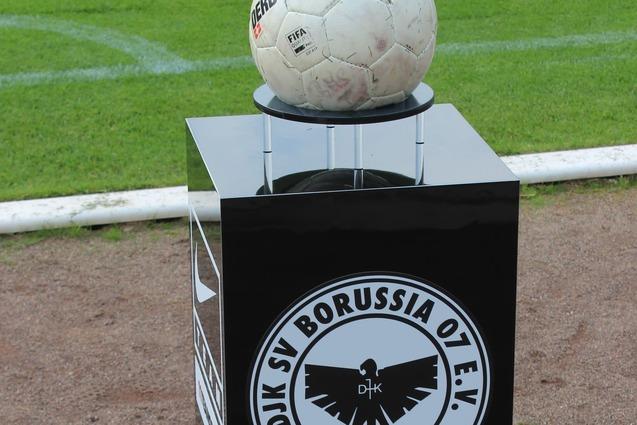 Ein Fussball auf einem schwarzen Podest mit dem Logo von DJK SV Borussia 07 e.V. M&uuml;nster im Hintergrund auf einem Rasenfeld.