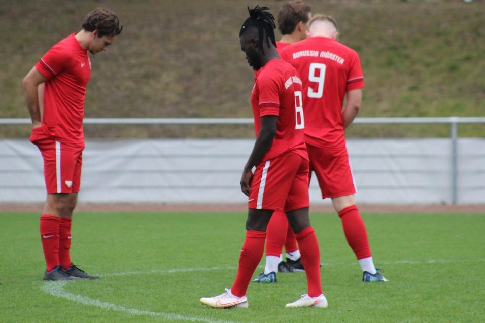 Drei Fu&szlig;ballspieler in roten Trikots stehen auf einem Rasenplatz, mit gesenkten K&ouml;pfen in Nachdenklichkeit.