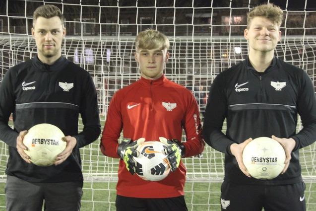 Drei Fu&szlig;ballspieler stehen vor einem Tor, zwei halten Fu&szlig;ball, einer tr&auml;gt Handschuhe und eine rote Jacke.