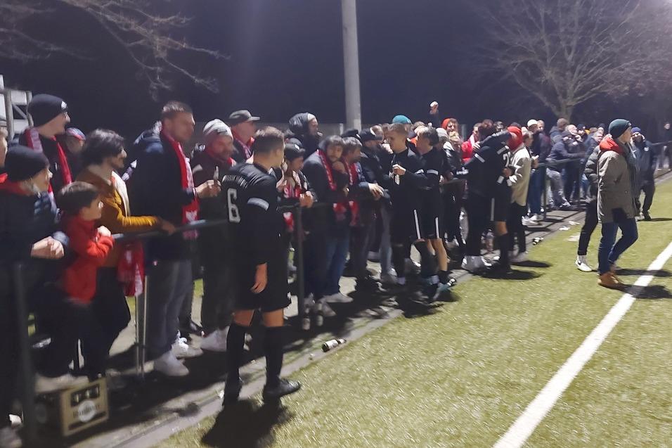 Gruppe von Zuschauern, darunter Kinder und Erwachsene, an einem Fu&szlig;ballfeld bei n&auml;chtlichem Licht.