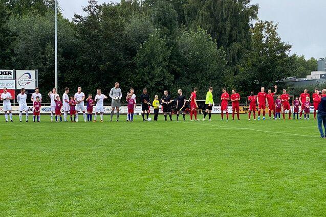 Zwei Fu&szlig;ballmannschaften stehen in einer Reihe auf einem gr&uuml;nen Rasenplatz, begleitet von einigen Zuschauern.