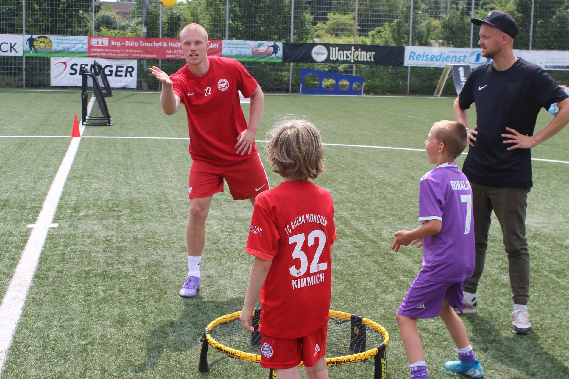 Trainer in rotem Outfit demonstriert Ballspiel mit zwei Kindern auf einem Fu&szlig;ballfeld. Kinder tragen Trikots und stehen um ein Ziel.