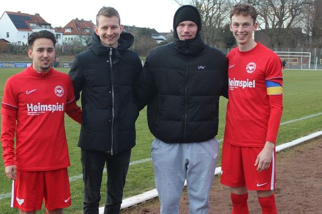 Vier M&auml;nner in Sportkleidung stehen auf einem Fu&szlig;ballfeld, zwei in roten Trikots, einer in einer Jacke, einer in grauen Jogginghosen.