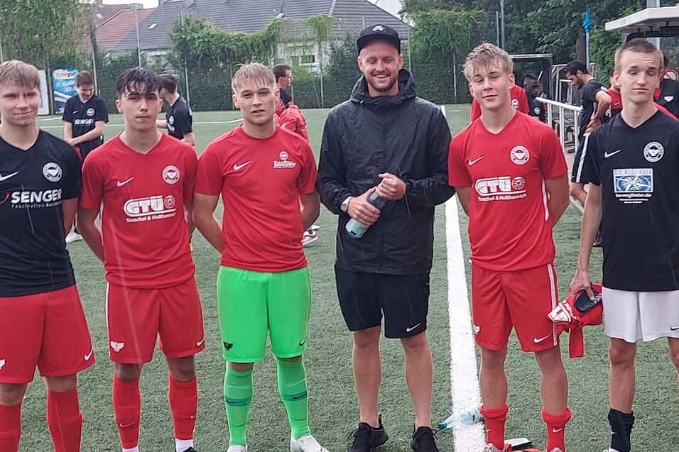 Sechs junge Fu&szlig;ballspieler in roten und schwarzen Trikots stehen auf einem Kunstrasenplatz, umgeben von Sporteinrichtungen.