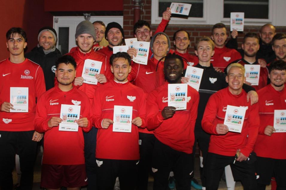 Gruppe von Fu&szlig;ballspielern in roten Trikots h&auml;lt Urkunden in der Hand, posierend vor einer roten Wand.