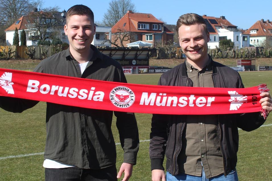 Zwei M&auml;nner halten einen roten Schal mit der Aufschrift &bdquo;Borussia M&uuml;nster&ldquo; auf einem Fu&szlig;ballfeld.
