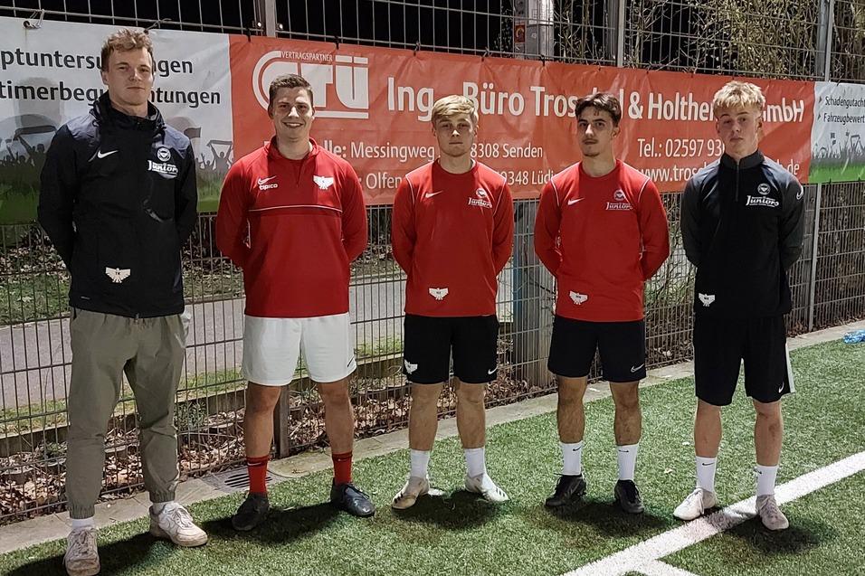 Vier Fu&szlig;ballspieler stehen neben einem Spielfeld, zwei in roten Trikots, einer in Schwarz und einer in sportlicher Kleidung.