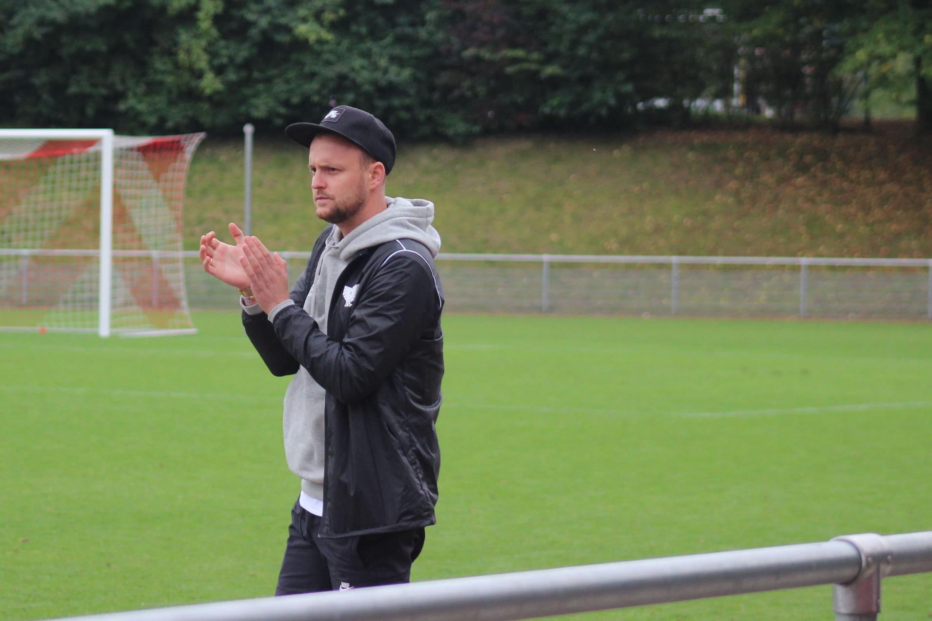 Mann im Sportoutfit klatscht am Rand eines Fu&szlig;ballfeldes, umgeben von gr&uuml;ner Wiese und B&auml;umen im Hintergrund.