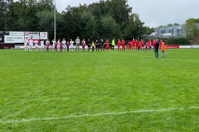 Zwei Fu&szlig;ballmannschaften stehen in einer Reihe auf einem gr&uuml;nen Rasenplatz, begleitet von einigen Zuschauern.