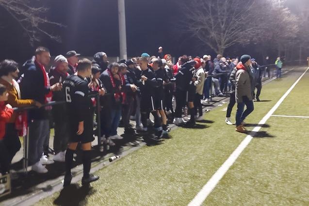 Gruppe von Zuschauern, darunter Kinder und Erwachsene, an einem Fu&szlig;ballfeld bei n&auml;chtlichem Licht.