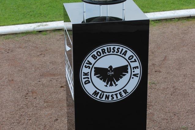 Ein Fussball auf einem schwarzen Podest mit dem Logo von DJK SV Borussia 07 e.V. M&uuml;nster im Hintergrund auf einem Rasenfeld.