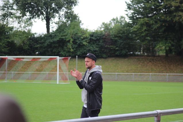Mann im Sportoutfit klatscht am Rand eines Fu&szlig;ballfeldes, umgeben von gr&uuml;ner Wiese und B&auml;umen im Hintergrund.