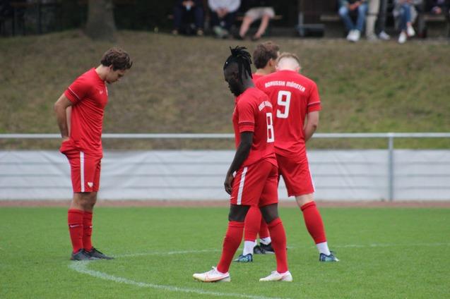Drei Fu&szlig;ballspieler in roten Trikots stehen auf einem Rasenplatz, mit gesenkten K&ouml;pfen in Nachdenklichkeit.