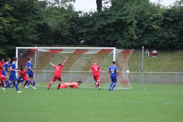 Fu&szlig;ballspieler in roten und blauen Trikots k&auml;mpfen um den Ball vor einem Tor, der Torwart liegt am Boden.