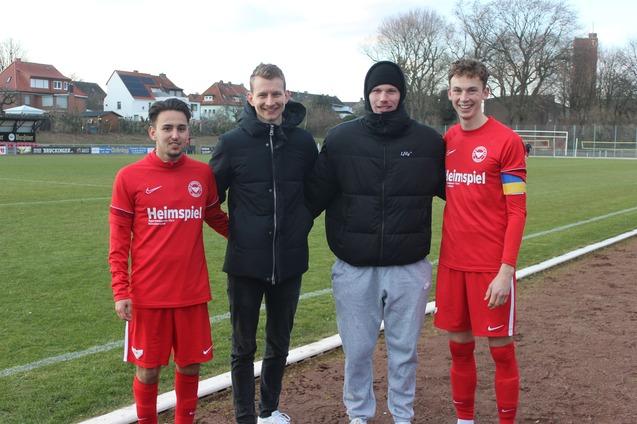 Vier M&auml;nner in Sportkleidung stehen auf einem Fu&szlig;ballfeld, zwei in roten Trikots, einer in einer Jacke, einer in grauen Jogginghosen.