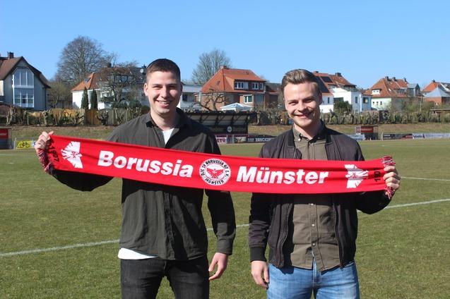 Zwei M&auml;nner halten einen roten Schal mit der Aufschrift &bdquo;Borussia M&uuml;nster&ldquo; auf einem Fu&szlig;ballfeld.