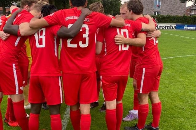 Mannschaft in roten Trikots umarmt sich vor einem Spiel auf einem Rasenplatz, im Hintergrund eine Sportanlage.