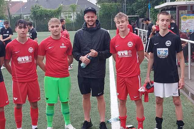 Sechs junge Fu&szlig;ballspieler in roten und schwarzen Trikots stehen auf einem Kunstrasenplatz, umgeben von Sporteinrichtungen.